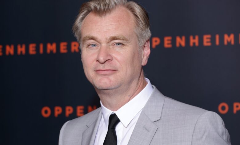 Christopher Nolan in a grey suit at a film event, with 'Oppenheimer' backdrop.
