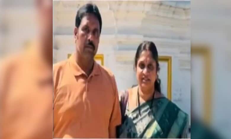 A man and woman standing together indoors, looking serious, with a blurred background.