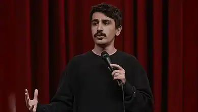 Turkish comedian holding microphone on stage with red curtain background.
