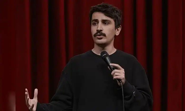 Turkish comedian holding microphone on stage with red curtain background.