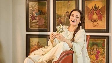 Dia Mirza smiling and holding a bouquet, seated in front of traditional Indian artwork.