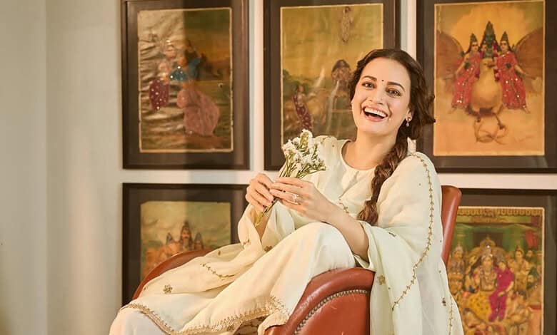 Dia Mirza smiling and holding a bouquet, seated in front of traditional Indian artwork.