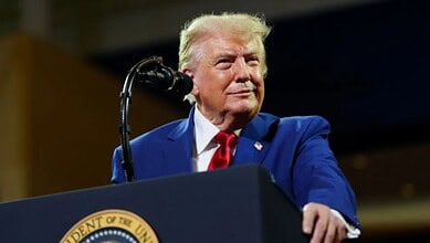 Donald Trump at a podium during a speech, wearing a blue suit and red tie, with a serious expression.