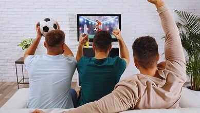 Three young men cheering and celebrating while watching a football game on TV.
