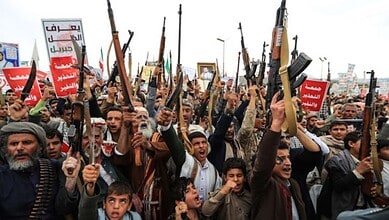 Houthi fighters protest holding rifles and banners in a large gathering.