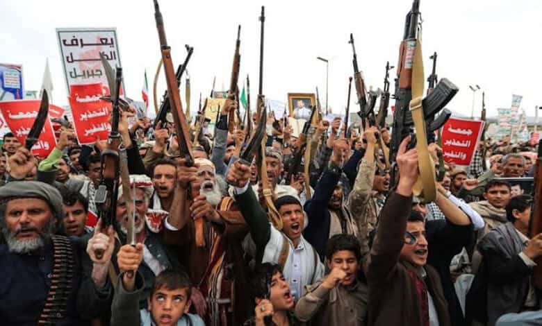 Houthi fighters protest holding rifles and banners in a large gathering.
