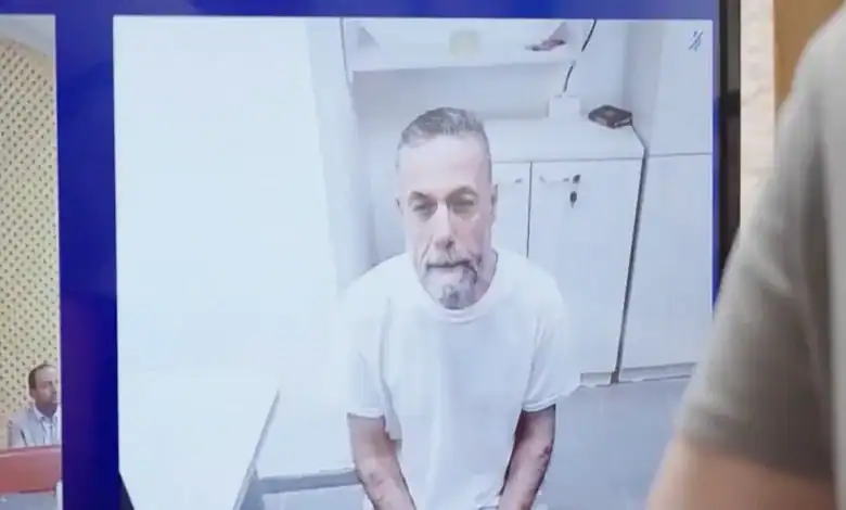 Hussam Abu Safiya appears on a courtroom video screen during a remote hearing, seated in a detention room wearing a white T-shirt.