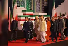 Indian delegation in formal attire pays tribute at a memorial event with Iranian flags in the background.