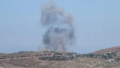 Large explosion with thick smoke rising over a hillside, with buildings and sparse vegetation.