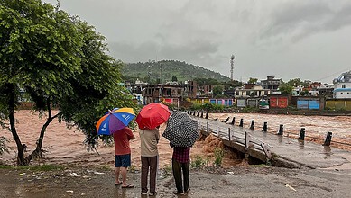 People with umbrellas standing near a flooded road during heavy rains.