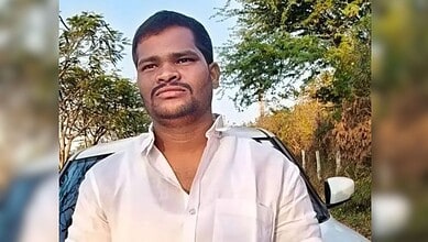 Man in white shirt standing outdoors with trees and a car in the background.