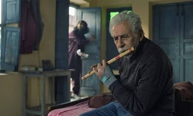 Elderly man playing flute indoors with a woman in the background at an open window.