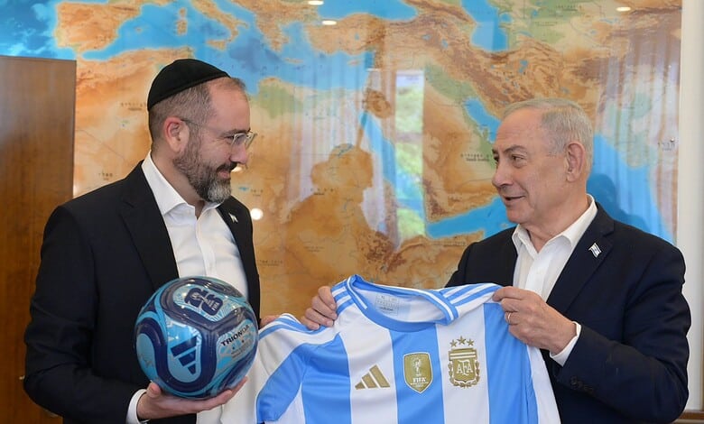 Benjamin Netanyahu and Argentine official holding a football and a jersey in front of a world map.