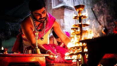 A Hindu priest in traditional attire performs an evening puja ritual, lighting a multi-tiered brass diya lamp beside a temple altar.