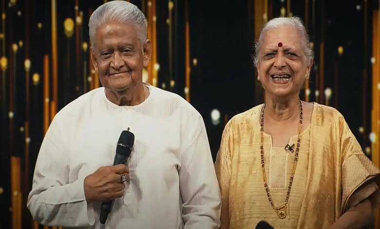 Elderly man and woman smiling at an event, woman holding a shawl, in a festive setting.