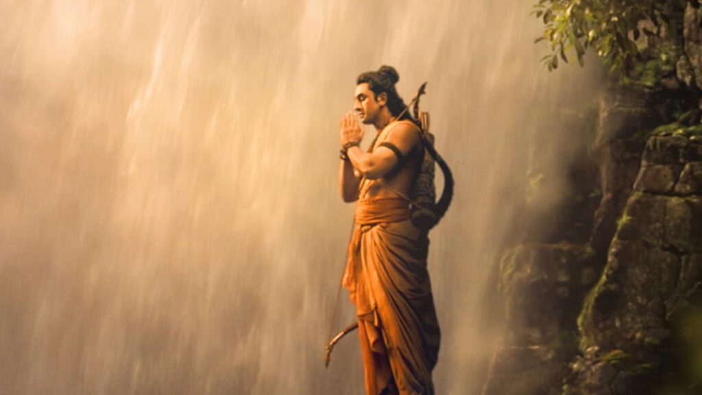 A man dressed as Lord Rama praying near a waterfall in a forest setting.