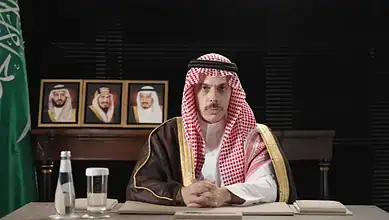 Saudi Foreign Minister in a formal meeting, wearing traditional attire, with Saudi flags in the backdrop.