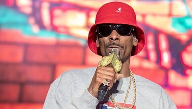 Snoop Dogg wearing a red hat and sunglasses, holding a microphone in front of a colourful background.