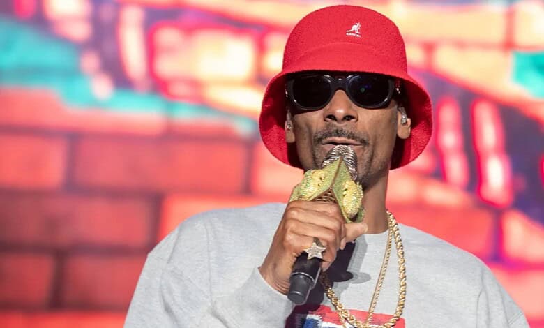 Snoop Dogg wearing a red hat and sunglasses, holding a microphone in front of a colourful background.