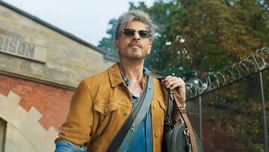 A man with grey hair and sunglasses wearing a brown jacket and blue shirt outdoors.