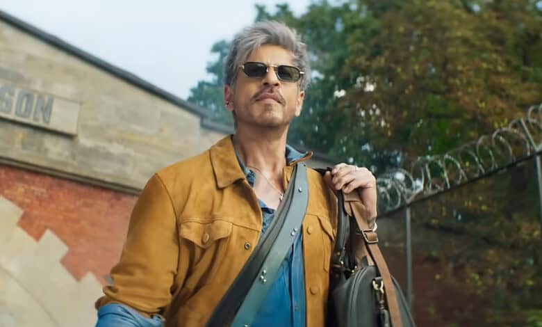 A man with grey hair and sunglasses wearing a brown jacket and blue shirt outdoors.
