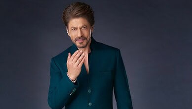 Shah Rukh Khan in a dark teal suit, posing thoughtfully against a dark background.