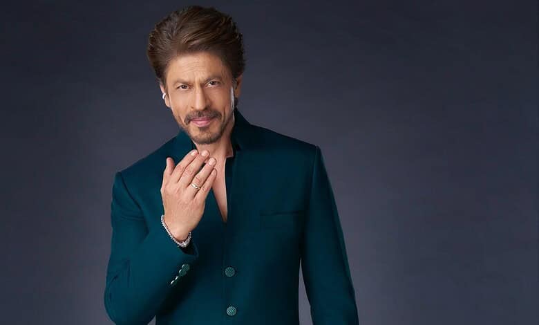 Shah Rukh Khan in a dark teal suit, posing thoughtfully against a dark background.