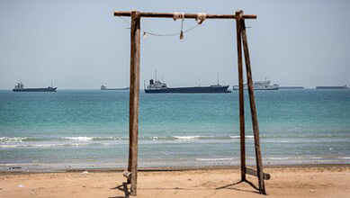 Empty wooden swing on sandy beach with ships in the distance.