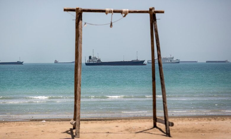 Empty wooden swing on sandy beach with ships in the distance.