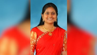 Smiling young woman in traditional red saree with gold jewellery against blue background.