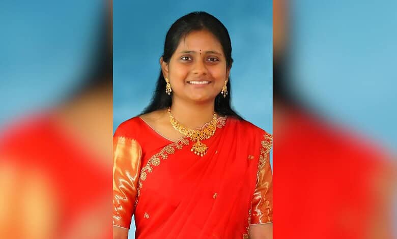 Smiling young woman in traditional red saree with gold jewellery against blue background.