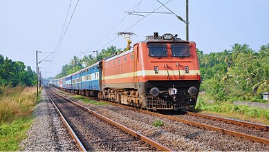 Indian Railways electric locomotive pulling passenger coaches through lush green countryside under overhead power lines.