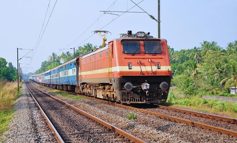 Indian Railways electric locomotive pulling passenger coaches through lush green countryside under overhead power lines.
