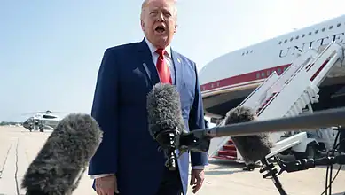 Donald Trump speaking at an airport with microphones in front, aircraft in background.