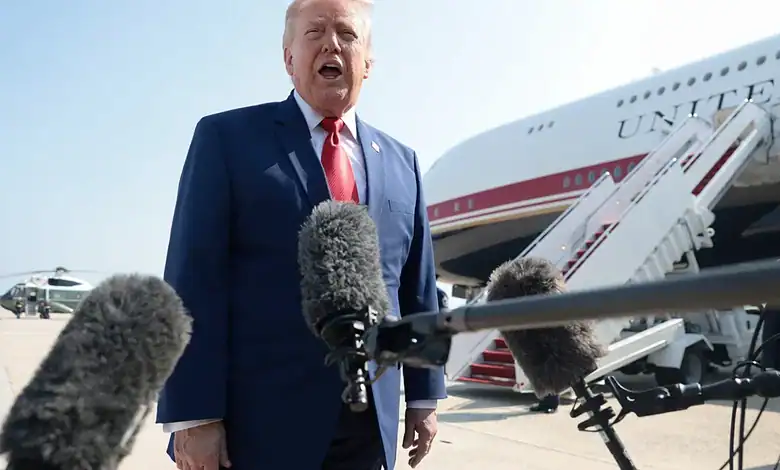 Donald Trump speaking at an airport with microphones in front, aircraft in background.
