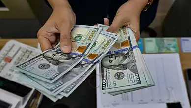 Person holding a stack of US $100 bills, close-up of cash and documents on table.