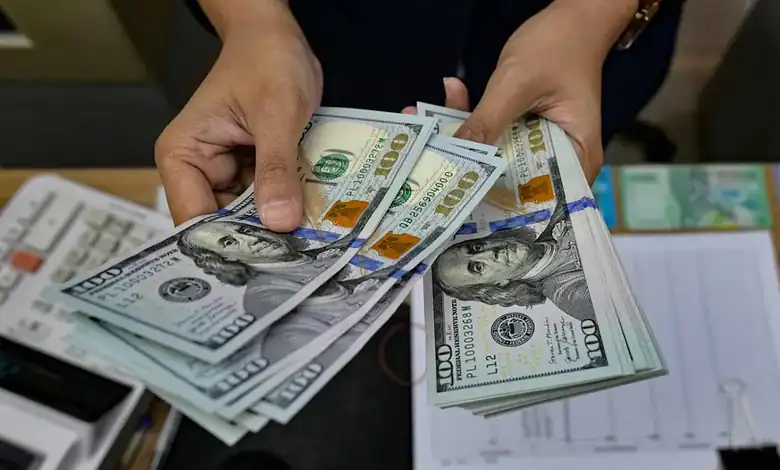 Person holding a stack of US $100 bills, close-up of cash and documents on table.