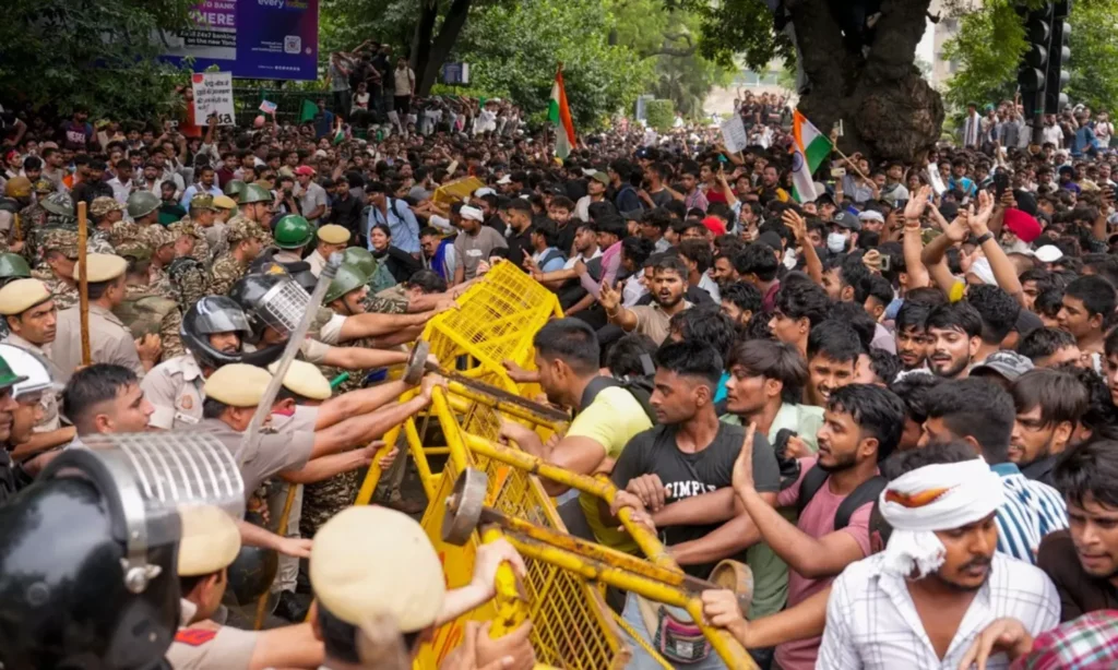 Large crowd protesting with police using shields and barricades in a tense standoff.