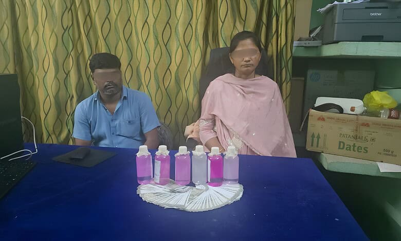 Two individuals seated at a table with pink bottles and a fan in front of them.