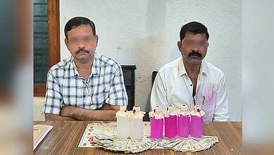Two men seated at a table with pink bottles, cash, and a cake, in an indoor setting.
