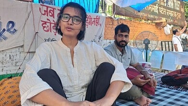 Young woman with glasses sitting outdoors during daytime, with a man in the background.
