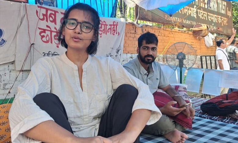 Young woman with glasses sitting outdoors during daytime, with a man in the background.