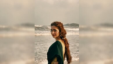 A woman with long, wavy hair in a green dress stands on the beach with waves behind her.
