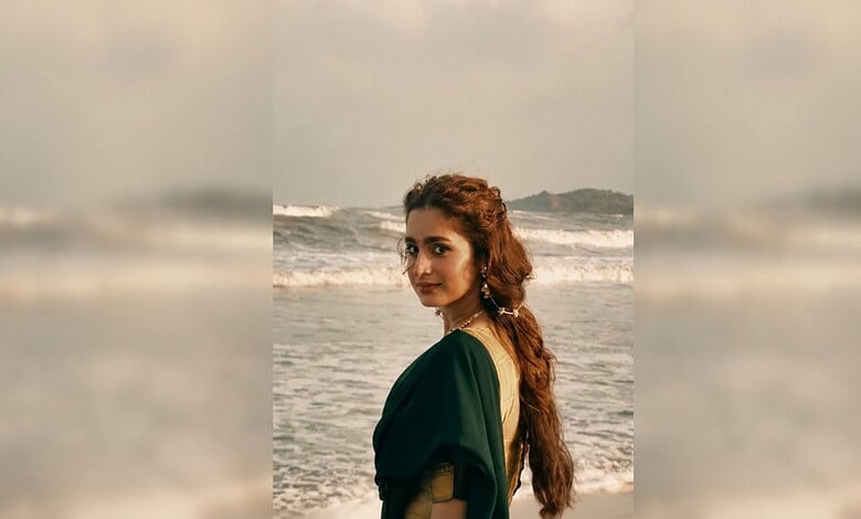 A woman with long, wavy hair in a green dress stands on the beach with waves behind her.