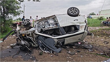 Upside-down car crash scene with debris and onlookers in the background.