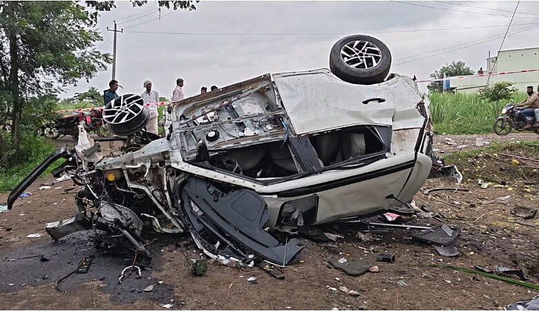 Upside-down car crash scene with debris and onlookers in the background.