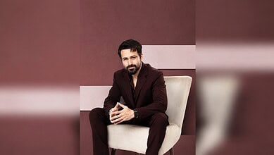 Emraan Hashmi sitting on a beige chair, wearing a dark suit, with a confident expression.