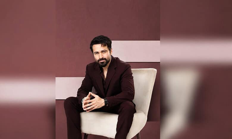 Emraan Hashmi sitting on a beige chair, wearing a dark suit, with a confident expression.