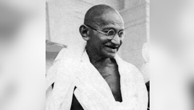 Black and white photo of Mahatma Gandhi smiling, wearing glasses and traditional attire.