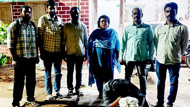 Group of six people standing outdoors with a woman in the centre, in front of a brick wall and plants.